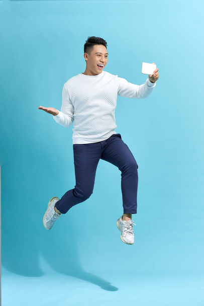 Портрет счастливого мужчины, прыгающего и кричащего, делая селфи на смартфоне на синем фоне - Фото, изображение
