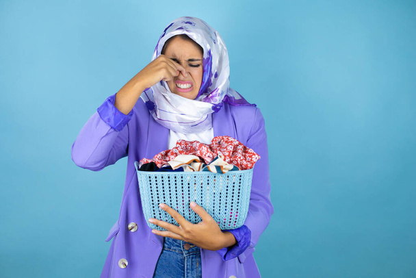 Giovane bella araba donna che indossa hijab islamico su isolato sfondo blu facendo lavori di casa in possesso di un cestino puzzolente con vestiti - Foto, immagini