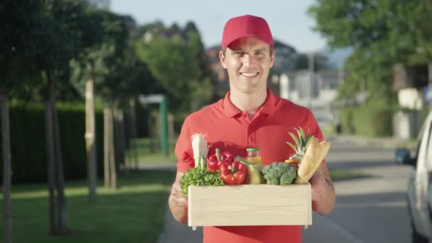 PORTRAIT: Cheerful delivery man holds a wooden box full of colorful groceries. - Footage, Video