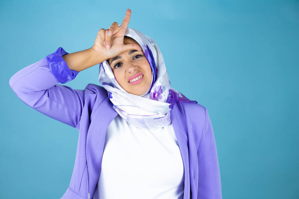 Jovem bela mulher árabe vestindo hijab islâmico sobre fundo azul isolado zombando de pessoas com dedos na testa fazendo gesto perdedor zombando e insultando. - Foto, Imagem