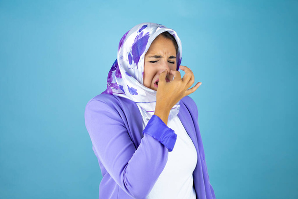 Giovane bella donna araba che indossa hijab islamico su sfondo blu isolato che odora di qualcosa di puzzolente e disgustoso, odore intollerabile, trattenendo il respiro con le dita sul naso - Foto, immagini