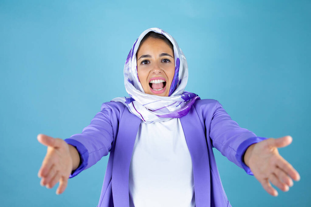 Young beautiful arab woman wearing islamic hijab over isolated blue background looking at the camera smiling with open arms for hug - Foto, afbeelding