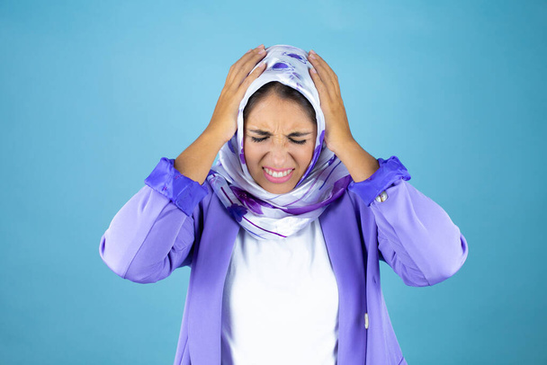 Joven mujer árabe hermosa usando hiyab islámico sobre fondo azul aislado que sufre de dolor de cabeza desesperado y estresado porque el dolor y la migraña con las manos en la cabeza - Foto, Imagen