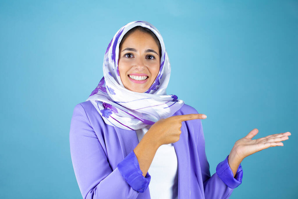 Молода красива арабська жінка в ісламському хіджабі над ізольованим синім тлом посміхається, показує і вказує щось, що на її руці
 - Фото, зображення