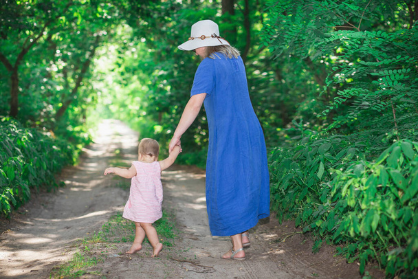 Η μαμά με το φόρεμα και το καπέλο μαθαίνει σε μια μικρή κόρη να περπατάει στο δάσος. - Φωτογραφία, εικόνα