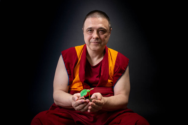 Monje budista en kesa roja. Un monje sosteniendo una planta verde en su mano - Foto, Imagen