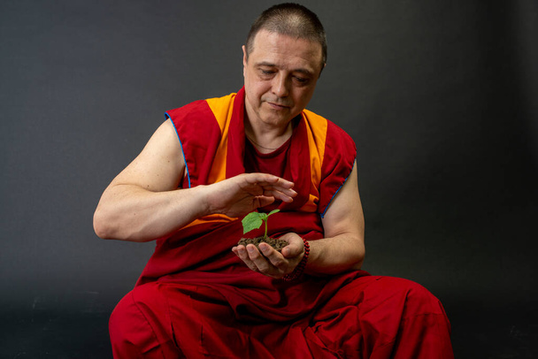 Буддийский монах в красной кесе. Монах держит в руке зеленое растение. - Фото, изображение