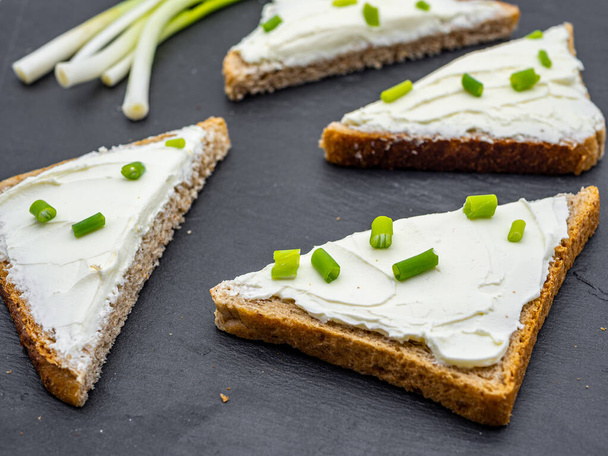 φέτες ψωμιού με κρέμα τυριού και φρέσκο πράσινο κρεμμύδι σε μαύρο σχιστόλιθο πρωινό παράγκα έννοια - Φωτογραφία, εικόνα