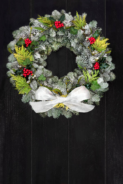 冬のベリーホリーと伝統的なクリスマスモミの花輪&素朴な木製のフロントドアの背景に白い弓と緑。冬至、クリスマス&新年の休暇シーズンのためのお祭りの組成。スペースのコピー. - 写真・画像