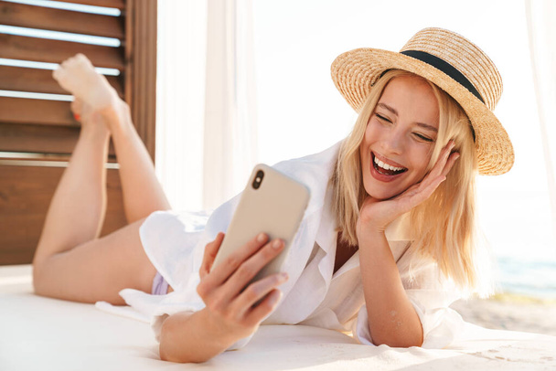 Image of laughing blonde woman laughing and taking selfie on cellphone while lying on bed at hotel - Foto, Imagen