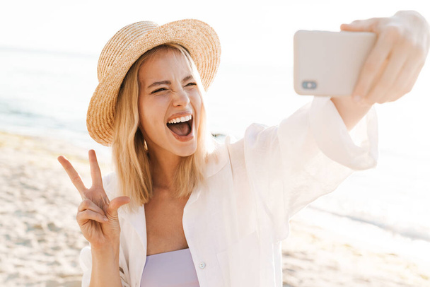 Image of excited woman showing peace sign and taking selfie on cellphone while standing at summer beach - Zdjęcie, obraz