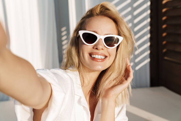 Image of seductive happy woman in sunglasses smiling while taking selfie photo at hotel on summer beach - Фото, зображення