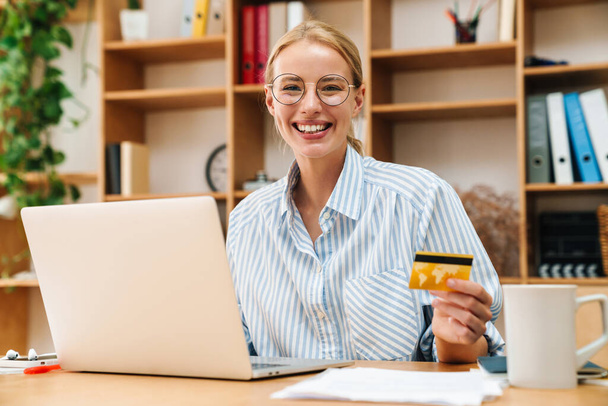 Image of joyful blonde woman holding credit card and smiling while working with laptop at table in office - Fotó, kép