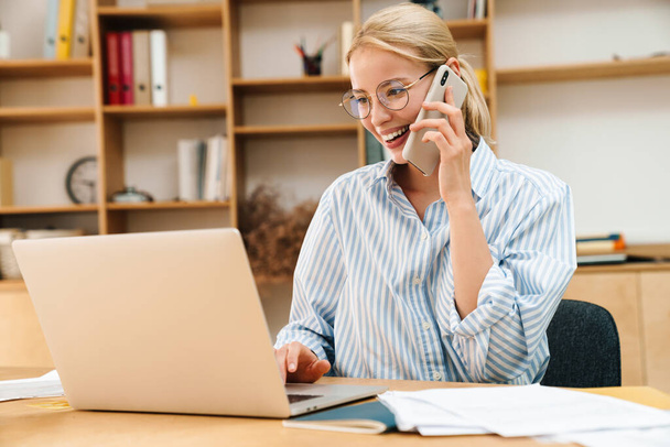 Image of joyful attractive businesswoman talking on cellphone while working with laptop at table in office - Фото, изображение