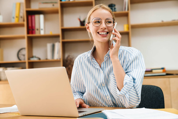 Image of joyful attractive businesswoman talking on cellphone while working with laptop at table in office - Photo, Image
