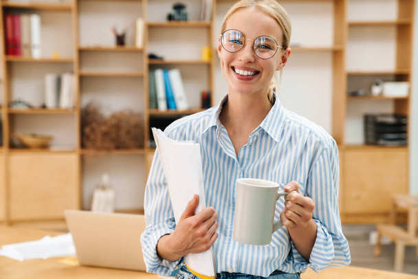 Image of joyful attractive woman in eyeglasses drinking coffee while posing with papers at office - Photo, Image