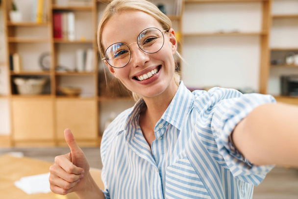 Image of cheerful attractive woman in eyeglasses showing thumb up while taking selfie photo at office - Fotografie, Obrázek
