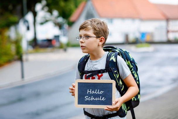 Rapazinho feliz com óculos e mochila ou mochila. Um estudante a caminho do liceu ou do liceu. Criança ao ar livre na rua. De volta à escola. Na mesa Olá escola em alemão. - Foto, Imagem