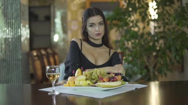 Молода спокуслива сексуальна брюнетка в чорній сукні з розщепленням їсть фрукти виноград один за столом у вишуканому ресторані
 - Кадри, відео