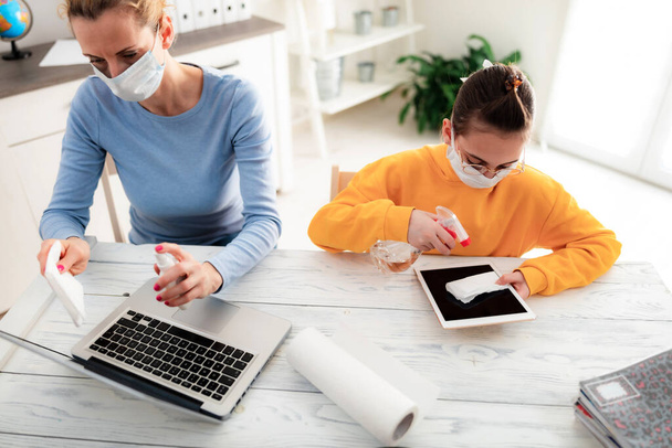 Sterilizing tablet and laptop with sanitizer. Child at home studying education, homeschooling, with private tutor / mother with protective mask in the time of viruses, flu and pandemic. - Photo, Image