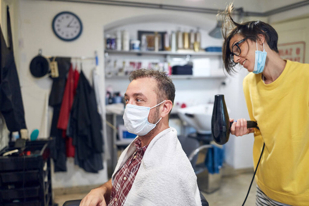 Peluquería y cliente en un salón con máscaras médicas durante la pandemia de virus. Trabajar con máscara de seguridad. - Foto, imagen
