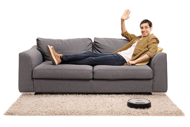 Guy lying on a sofa and waving at the camera while a robot vacuum cleaner is cleaning the carpet isolated on white background - Фото, зображення