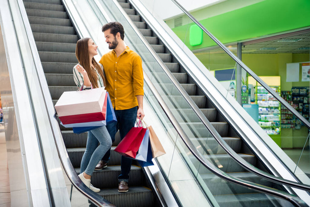 Full length photo of attractive lady handsome guy couple spend free time carry many bags moving up escalator shopping center hug look eyes wear casual jeans shirt outfit indoors - Φωτογραφία, εικόνα