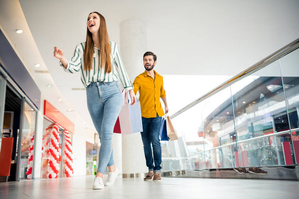 Low angle view full length photo of energetic woman see incredible 50 sales go walk hurry her husband follow hold many packages wear striped yellow shirt denim jeans in shopping mall center - Fotografie, Obrázek