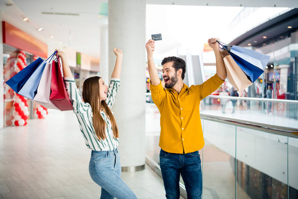 Photo of two people cheerful pretty lady handsome guy couple enjoy free time hold many bags walk shopping center raise hands use credit card discounts wear casual jeans shirt outfit indoors - Φωτογραφία, εικόνα
