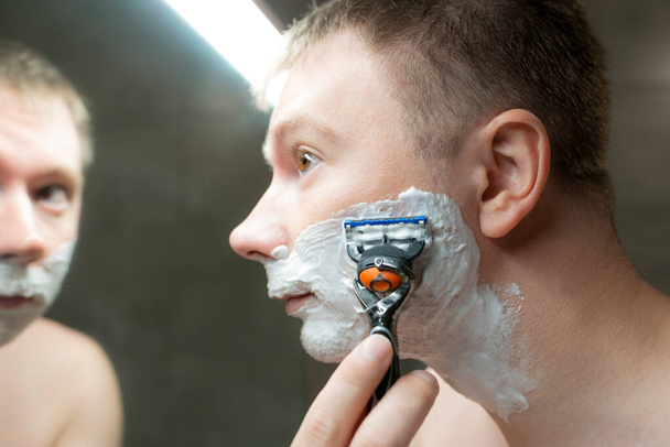 Guy shaves near the bathroom mirror. Holding a razor in hand - Photo, Image
