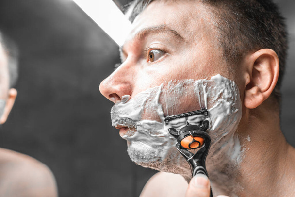O tipo faz a barba perto do espelho da casa de banho. Segurando uma navalha na mão - Foto, Imagem