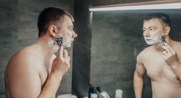 Ο τύπος ξυρίζεται κοντά στον καθρέφτη του μπάνιου. Κρατώντας ένα ξυράφι στο χέρι - Φωτογραφία, εικόνα