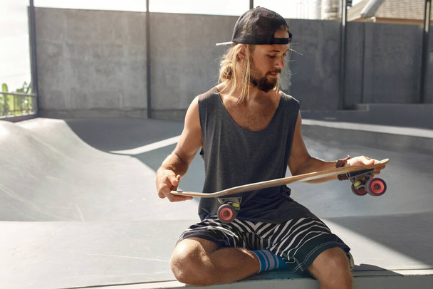 Skater Man At Skatepark. Portrait Of Handsome Guy In Casual Outfit With Skateboard Sitting On Concrete Ramp. Hipster With Extreme Sport Equipment At Park. Skateboarding As Urban Lifestyle. - Foto, imagen