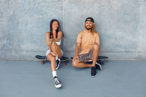 Skateboarding. Couple In Casual Outfit Sitting On Skateboards Against Concrete Wall At Skatepark. Urban Sport As Hobby Of Active Guys And Girls. Skater Subculture And Summer Activity As Lifestyle. - Фото, изображение