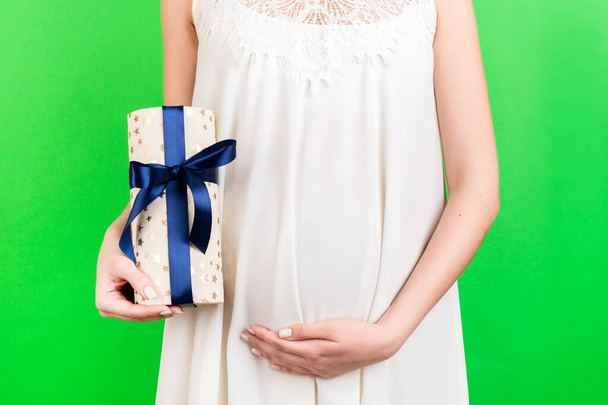 Cropped image of pregnant woman in white dress holding a gift box and touching her belly at green background. Expecting a baby boy. Copy space. - Фото, зображення