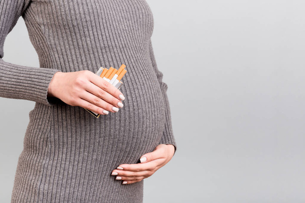 Zugeschnittenes Bild einer schwangeren Frau in grauem Kleid, die vor grauem Hintergrund eine Zigarettenschachtel hält. Das Rauchen während der Schwangerschaft ist verboten. Nikotinmissbrauch. - Foto, Bild