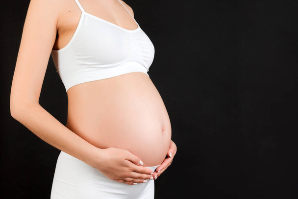 Cropped image of pregnant woman in white underwear embracing her big abdomen at black background. Maternity concept. Copy space. - Foto, afbeelding
