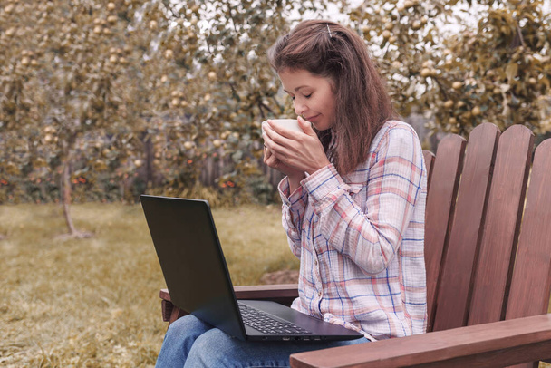 Молода жінка сидить на стільці в саду біля ноутбука з чашкою чаю в руках
 - Фото, зображення