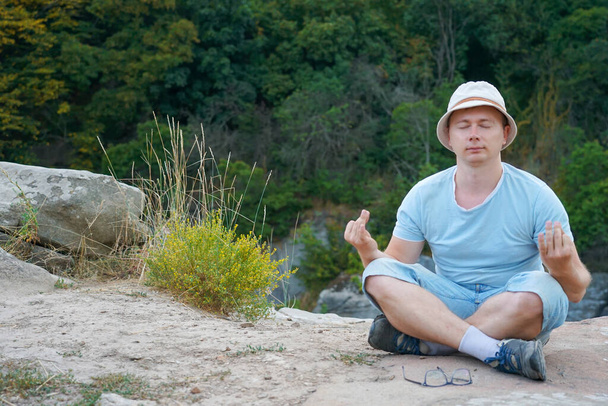 Um cara se senta em uma rocha nas montanhas em uma pedra, vestido com uma camiseta e shorts - Foto, Imagem