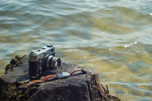 Вінтажний аналоговий фотоапарат і дивитися зі шкіряним ремінцем, що лежить на пеньку поруч з водою. Крупним планом
 - Фото, зображення