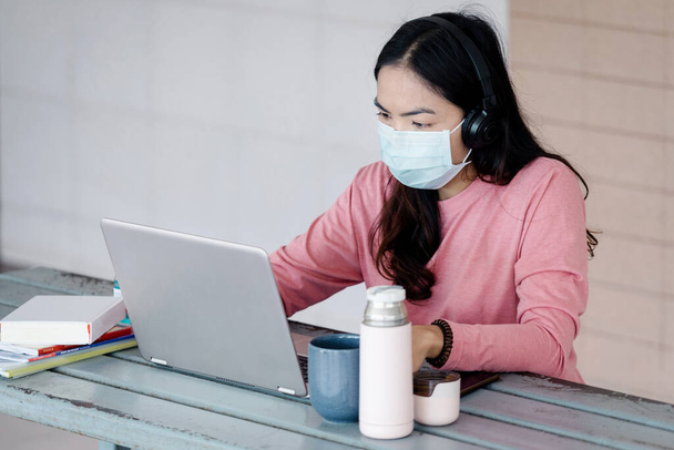Young Asian businesswoman with face mask working on laptop at home during Coronavirus or COVID-19  pandemic.. Work from home concept. Stock photo - Фото, изображение