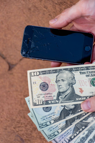 Сідней, Австралія - 2019-11-01 Рука, що тримає телефон з сильно розтрощеним екраном і доларами США, щоб заплатити за ремонт. Випадкова концепція. - Фото, зображення