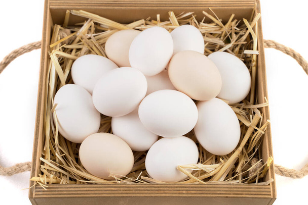 Белые и коричневые куриные яйца лежат на соломе в деревянной коробке. - Фото, изображение