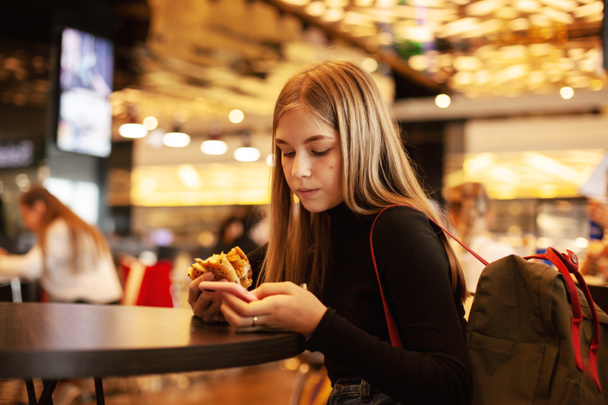 A beautiful teenage girl sitting in a cafe looks at the phone and eats a hamburger. Happy child eating junk food in a restaurant. Side view - Fotografie, Obrázek