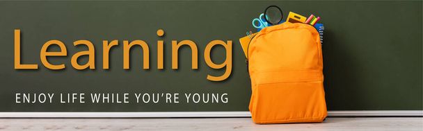horizontales Bild des gelben Rucksacks voller Schulpapier in der Nähe der grünen Tafel mit Lernen, genießen Sie das Leben, während Sie jung sind Schriftzug  - Foto, Bild