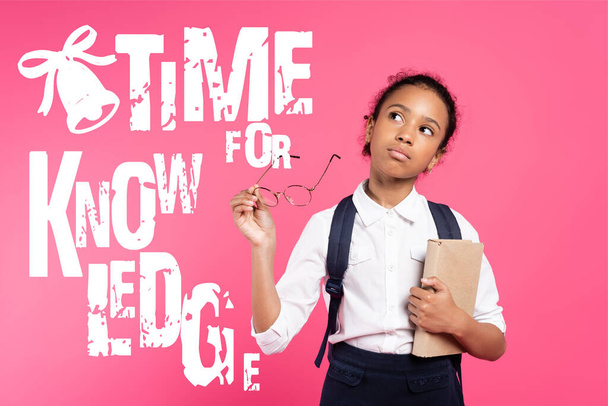 pensive african american schoolgirl with book and eyeglasses standing near time for knowledge lettering on pink - Foto, afbeelding