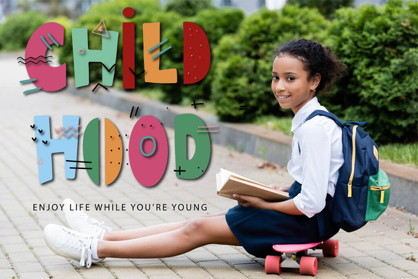 african american schoolgirl holding book while sitting on penny board near childhood, enjoy life while you are young lettering  - Photo, Image