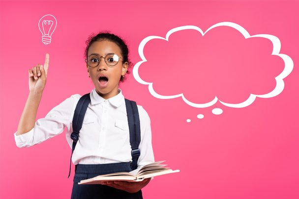 shocked african american schoolgirl in glasses holding book and showing idea gesture near thought bubble on pink - Photo, Image