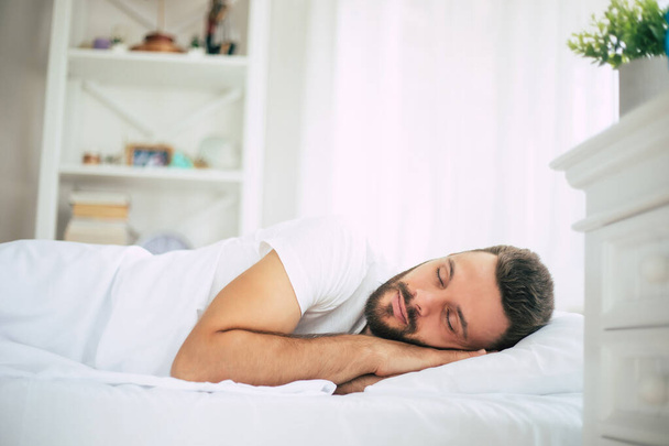 Handsome young bearded man is sleeping in a white big bed in a comfortable bedroom - Фото, изображение