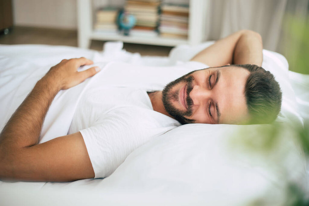 Handsome young bearded man is sleeping in a white big bed in a comfortable bedroom - Foto, Bild